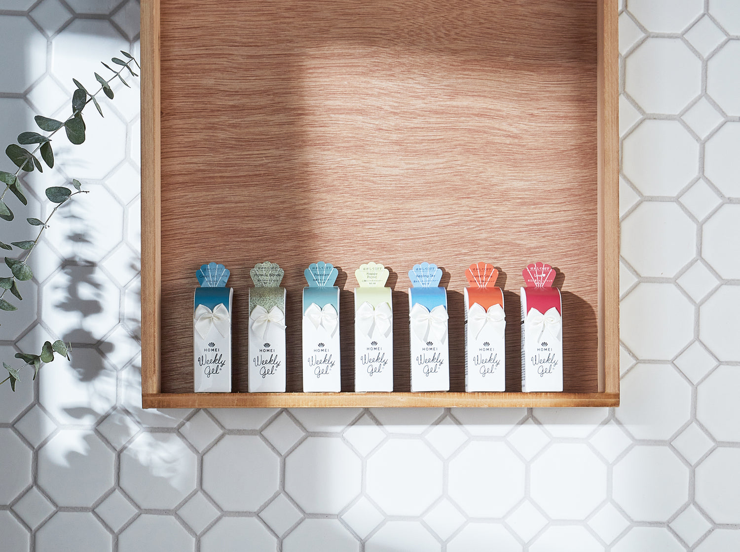 Colorful Weekly Gel product bottles are lining up on a hanging wood shelf. The background is white tiles. Natural light. 